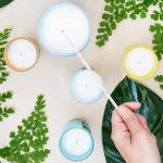 A hand lights a white tallow candle surrounded by several other candles in jars, green leaves, and fern sprigs on a light surface. Text at the bottom reads "Homemade Tallow Candles.