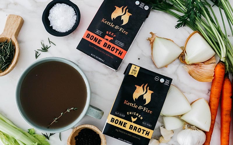 Two cartons of Kettle and Fire Bone Broth surrounded by vegetables and garnishes.