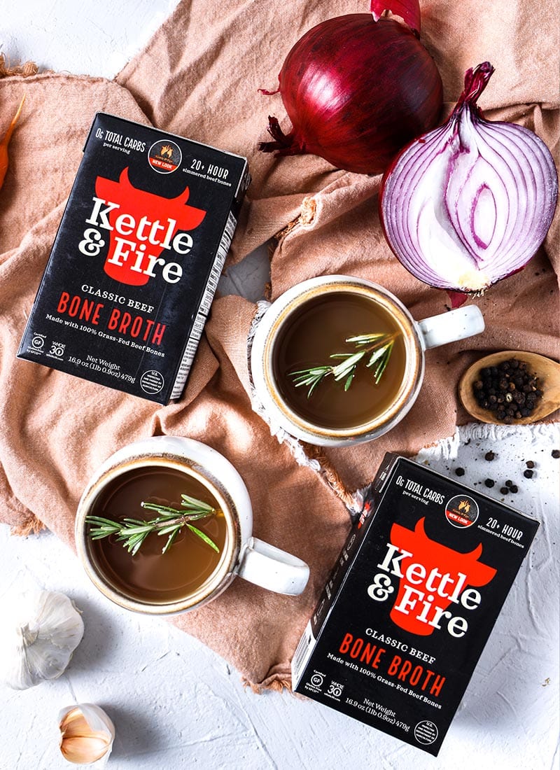 Two cartons of kettle & fire beef bone broth on a brown cloth surrounded by ingredients.