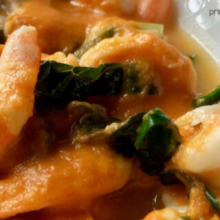 Close-up of shrimp in a rich, orange moqueca de camarao sauce with green leafy vegetables.