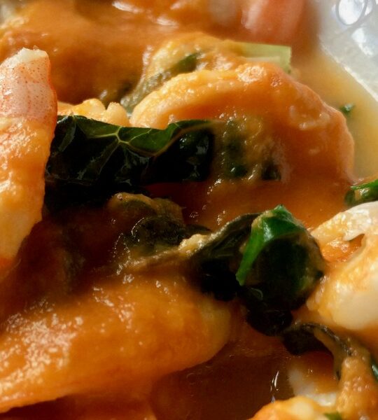 Close-up of shrimp in a rich, orange moqueca de camarao sauce with green leafy vegetables.