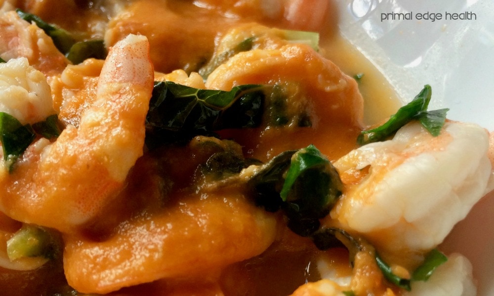 Close-up of shrimp in a rich, orange moqueca de camarao sauce with green leafy vegetables.