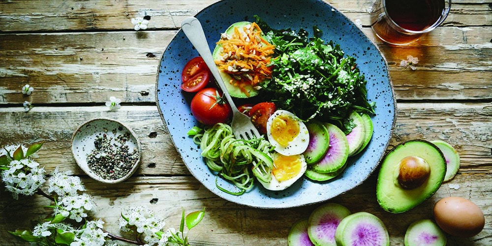 Vegetable keto salad in a blue plate with a fork surrounded by a glass with brown liquid, half avocado and an egg.