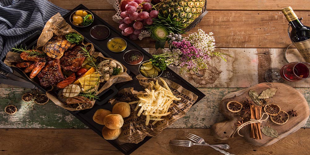 A big serving plate with grilled meat and shrimp and different sauces next to a wooden b oard with different spices.