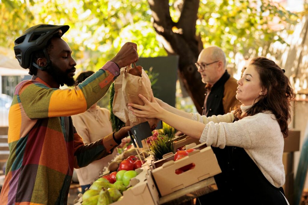 A woman is handing a man a box of vegetables from a farmers' market stand.
