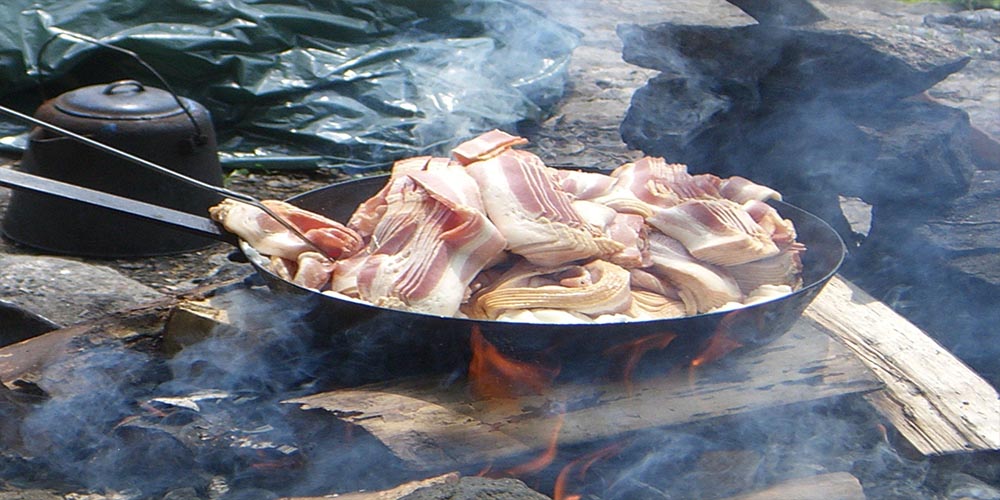 Stripes of bacon in a big pan being cooked using fire wood.