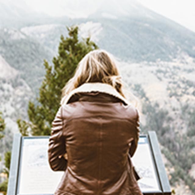 A woman in a brown jacket looking at a sign in front of a mountain.
