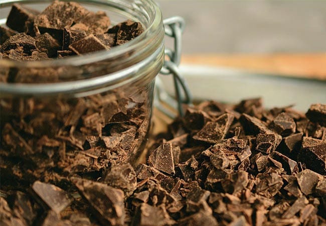 Chopped cacao paste for keto chocolate chip cookies in a glass jar surrounded by pieces of the same cacao paste.