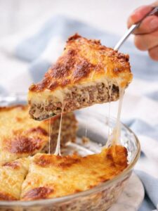 A slice of easy cheeseburger pie with a browned, melted cheese topping is being lifted from a glass dish, revealing gooey, stretchy cheese.