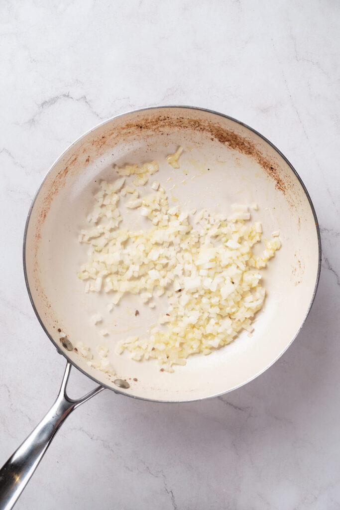 Chopped onions sautéing in a frying pan on a marble countertop.