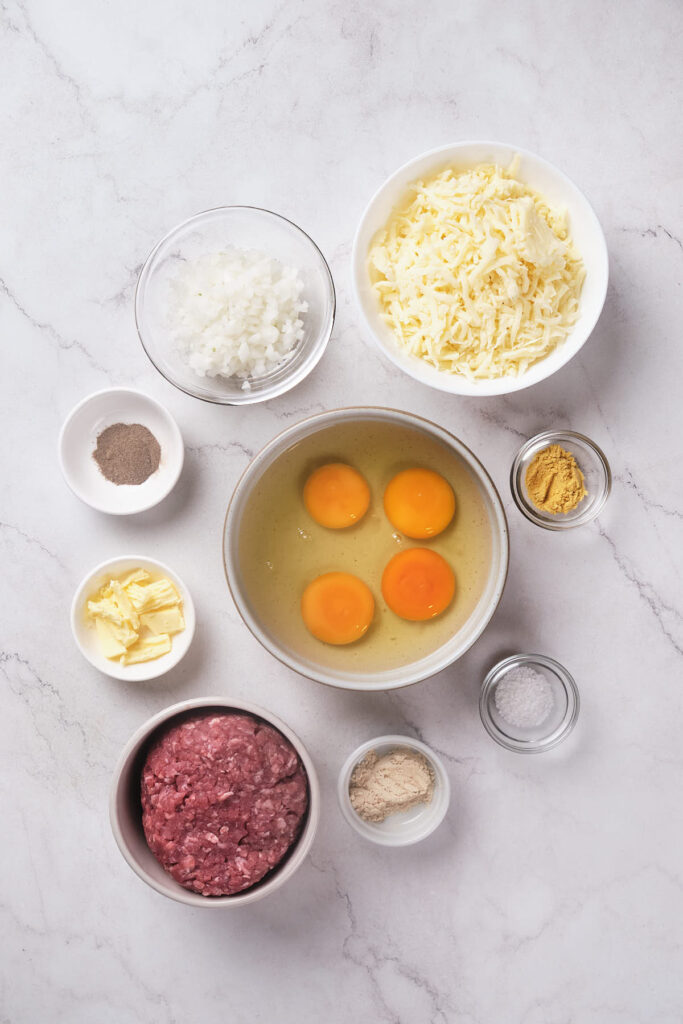 Various ingredients arranged on a marble surface, including eggs, ground meat, cheese, onions, and spices.