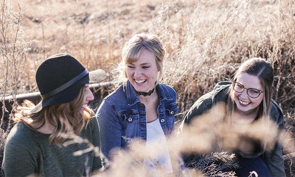 Three women laughing in a field of tall grass.