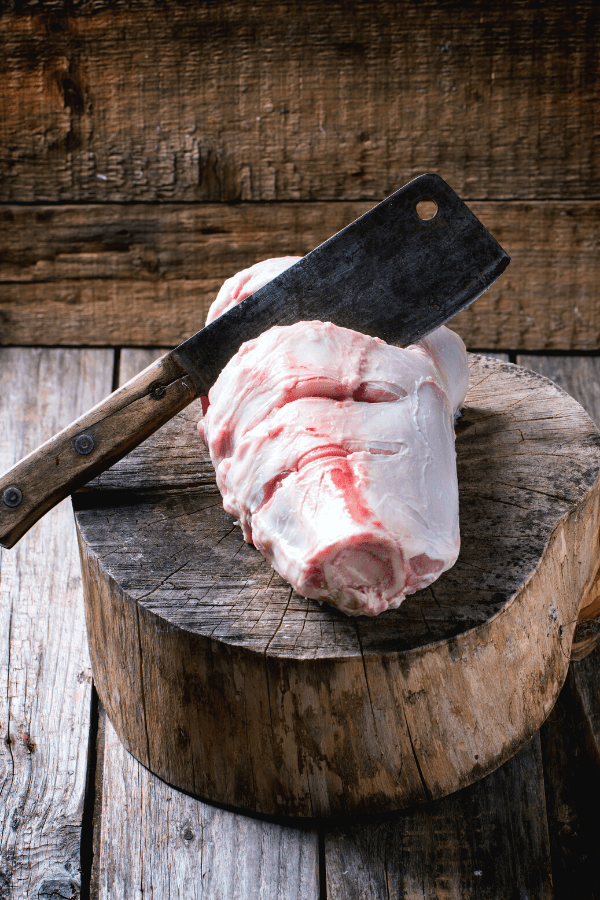 Butchers cleaver cutting into uncooked beef shank on wood stump.
