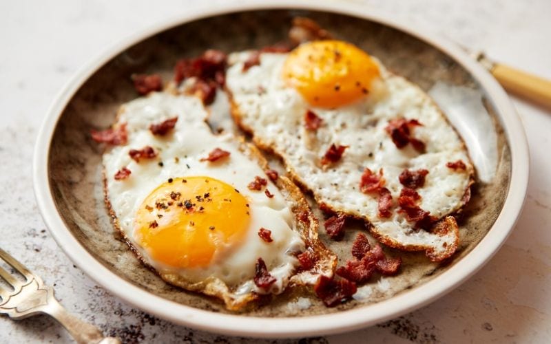Two fried eggs on a plate, accompanied by sizzling bacon.