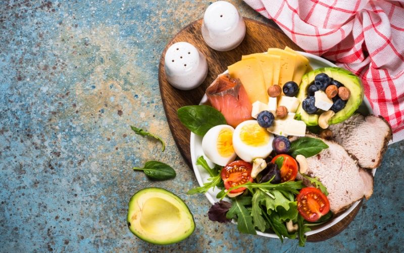 A keto-friendly salad with meat, eggs, vegetables and fruit on a blue background.