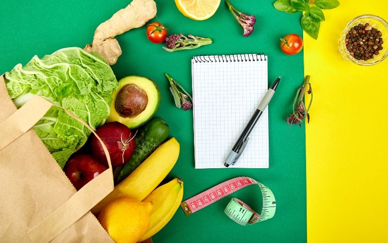 A keto-friendly paper bag filled with an assortment of fresh fruits and vegetables on a vibrant green and yellow background.