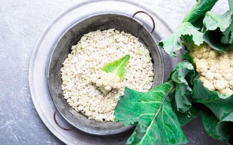 Uncooked cauliflower rice in a strainer next to fresh whole cauliflower.