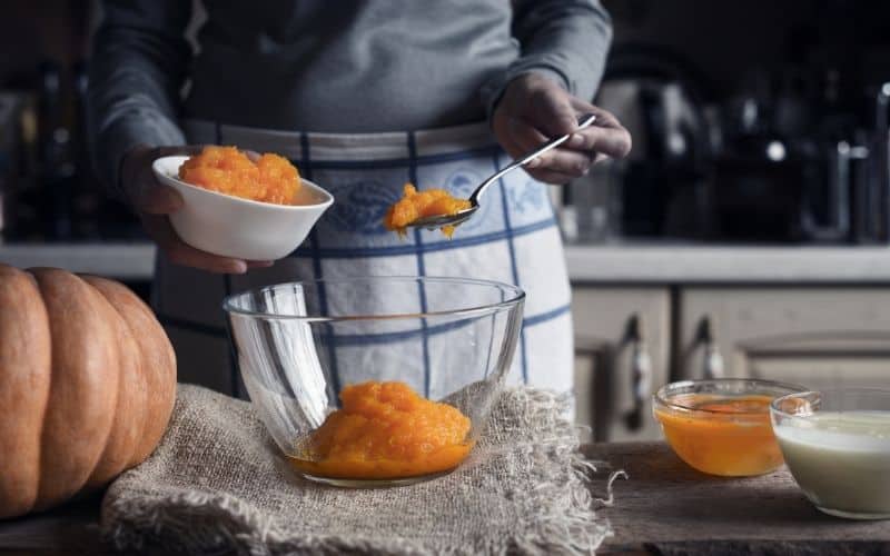 A person transferring an orange jam from small bowl to a large glass bowl.