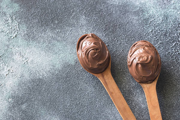 Two wooden spoons full of melted chocolate on a grey surface.