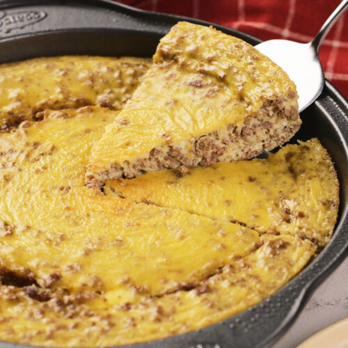 A metal pan with a baked carnivore casserole of egg and ground meat, with one slice being lifted out on a spatula; a red checkered cloth is in the background.
