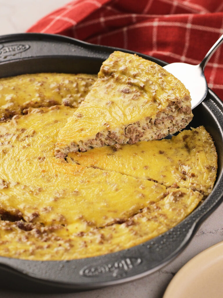 A metal pan with a baked carnivore casserole of egg and ground meat, with one slice being lifted out on a spatula; a red checkered cloth is in the background.