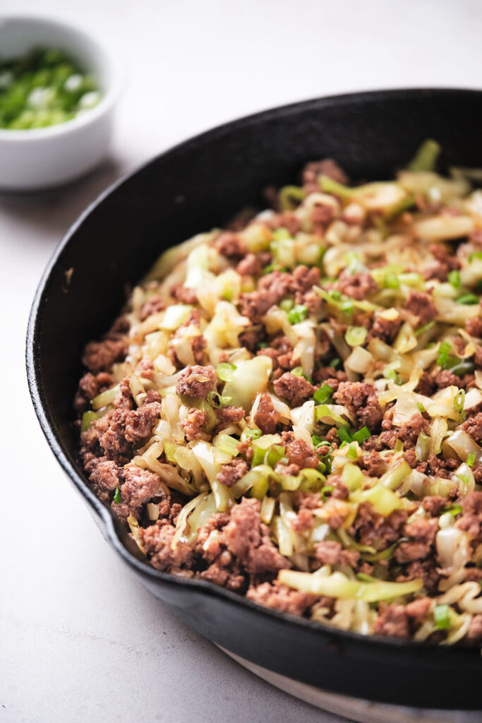 Cooked ground beef with chopped onions and cabbage in a black skillet, with a small bowl of chopped green onions in the background on a white surface.
