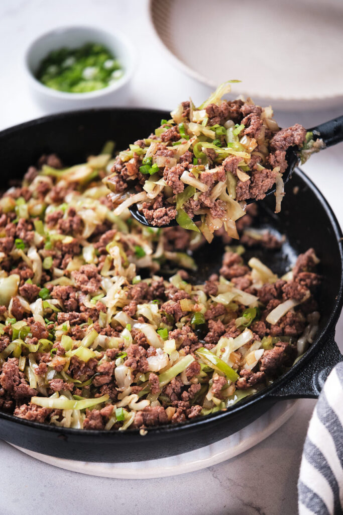 Cooked ground beef, chopped onions, and cabbage in a black skillet, with a spatula lifting some of the stir fry mixture, on a light-colored countertop.