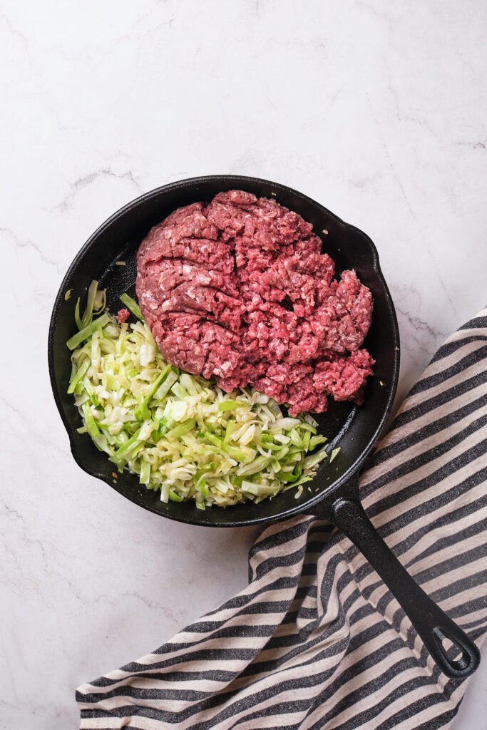 A cast-iron skillet containing raw ground beef and chopped cabbage.