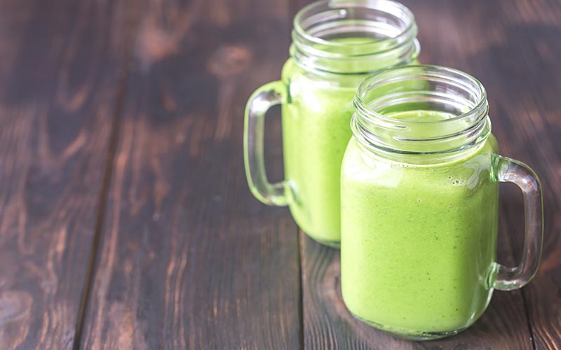 Two glasses of green smoothie on a brown surface.
