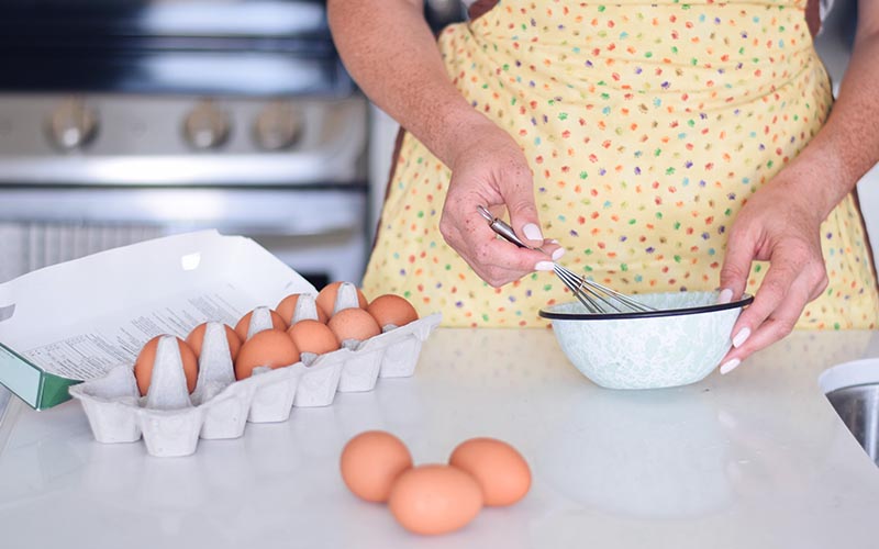 A person holding a wire whisk and a bowl with a tray of eggs.