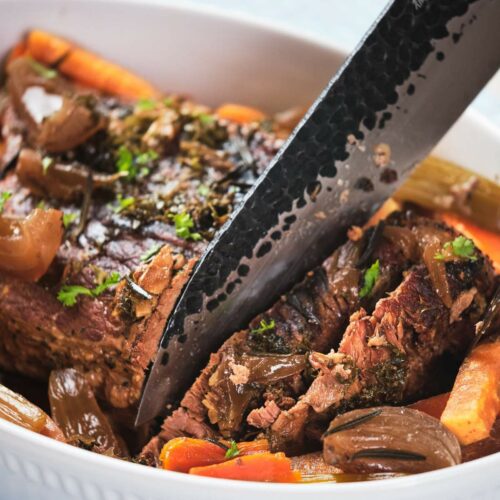A close-up of keto pot roast with carrots and onions in a white serving dish, garnished with herbs, with a knife resting on top.