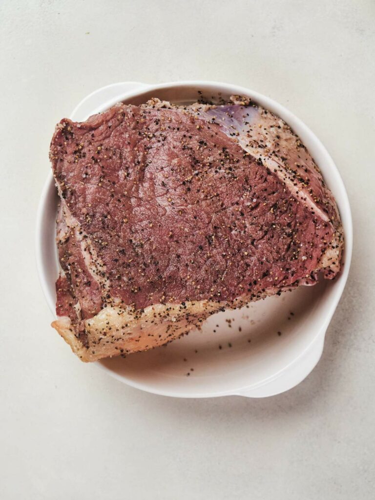 A raw steak seasoned with black pepper and salt on a white plate.