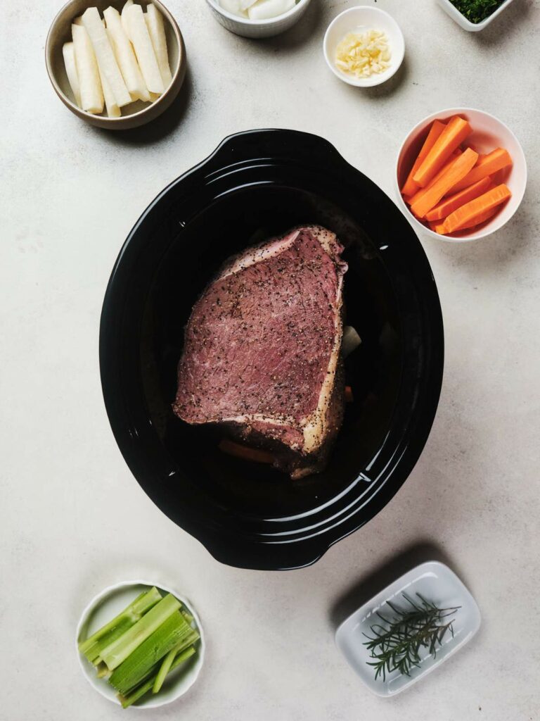 Piece of meat in a slow cooker with vegetables and separate small bowls of ingredients around it on a light surface.
