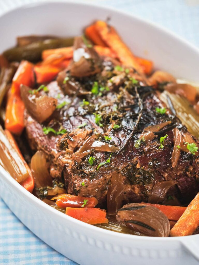 Pot roast with carrots, onions, and herbs in a white ceramic dish.