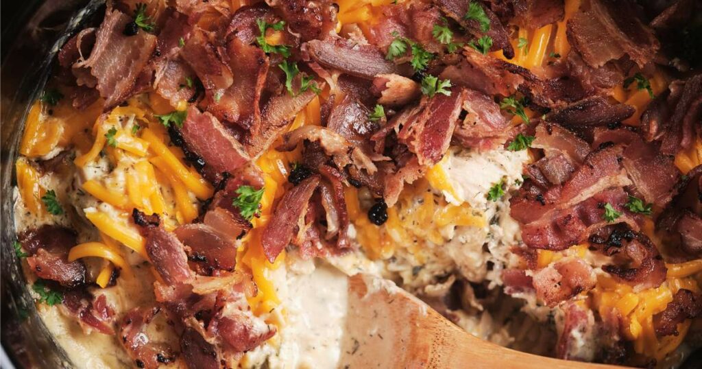 Close-up of a slow cooker with creamy crack chicken topped with crispy bacon bits and shredded cheddar cheese, garnished with parsley.