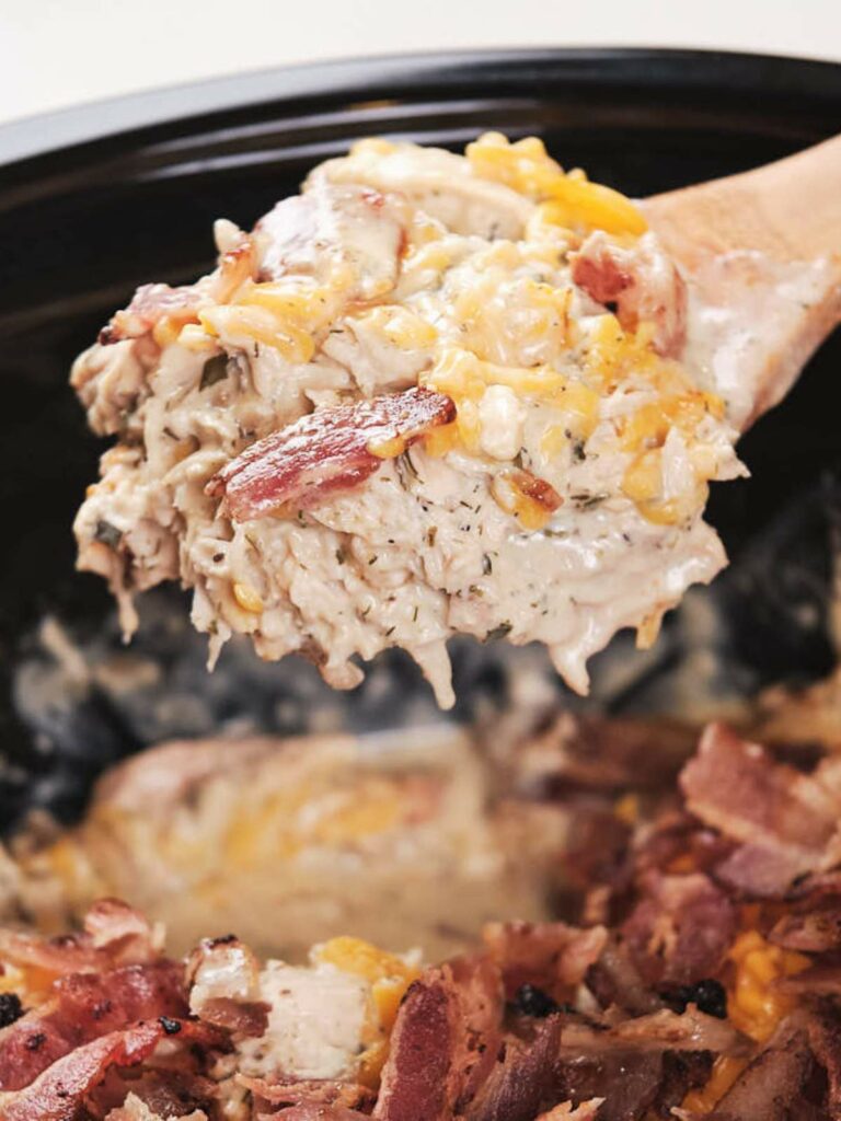 A close-up of a spoon lifting cheesy slow cooker crack chicken casserole with bacon bits from a black dish.