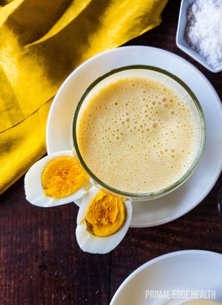 Egg smoothie in a glass on white saucer plate on a wooden surface.