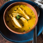 Yellow soup with meat in a bowl with spoon on blue plate.