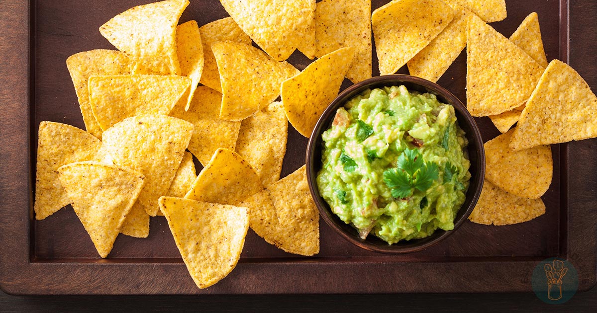 Tortilla chips arranged on a wooden board around a bowl of guacamole.