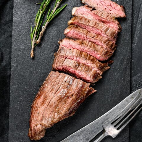 Sliced steak on a black slate board with herbs and a fork and knife.