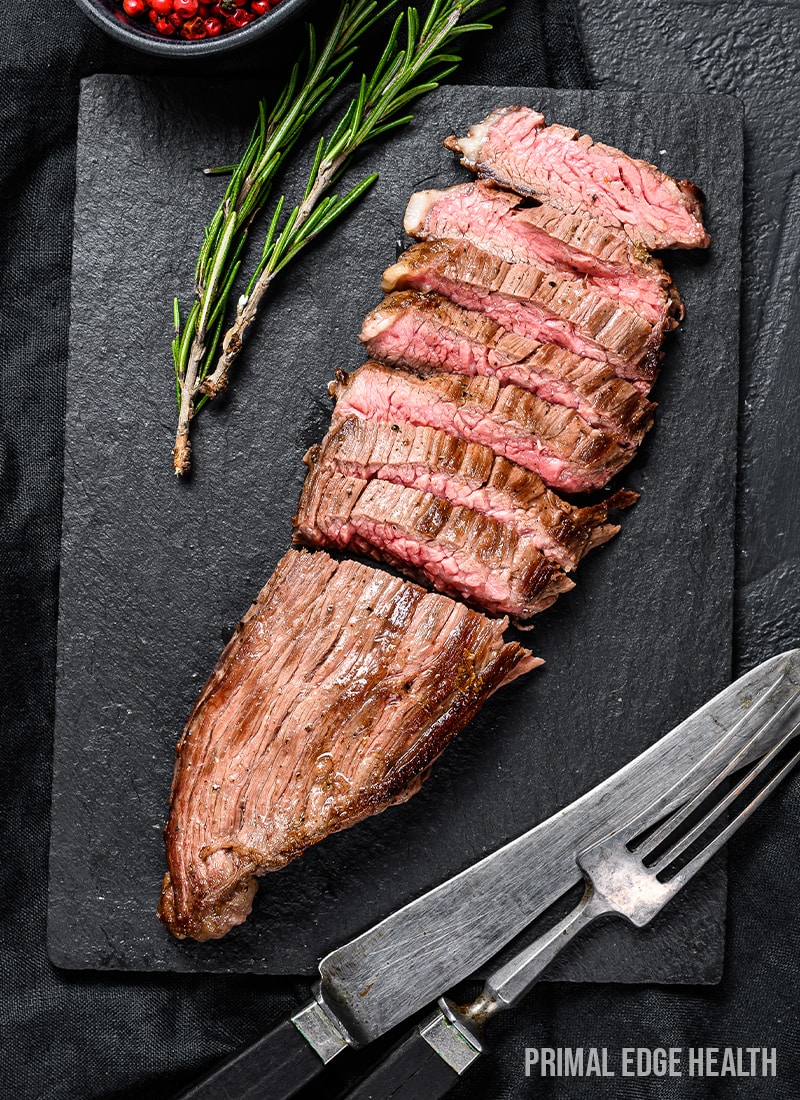 Sliced steak on a black slate board with herbs and a fork and knife.