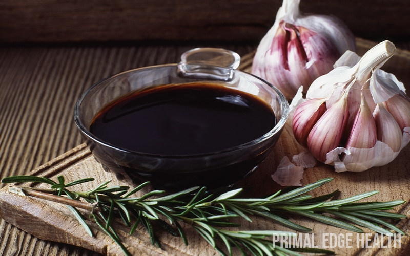 Bowl of black sauce with garlic and rosemary on wooden surface.