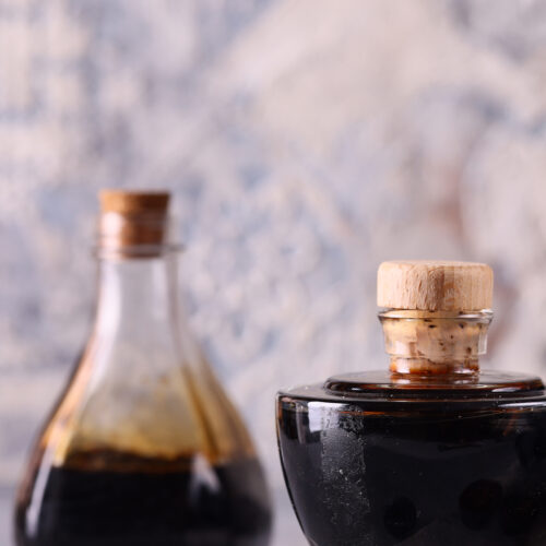 A glass jar with a cork stopper containing dark liquid, with a similar bottle blurred in the background.