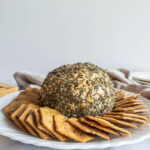 Herb coated cheese ball on white serving plate surrounded by crackers.