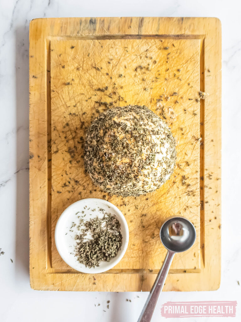 keto cheese ball on a wooden cutting board being rolled in herb mixture