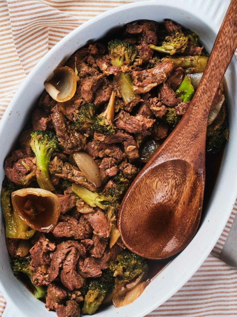 A keto dish of stir-fried beef and broccoli served in a white oval casserole with a wooden spoon, placed on a striped cloth.