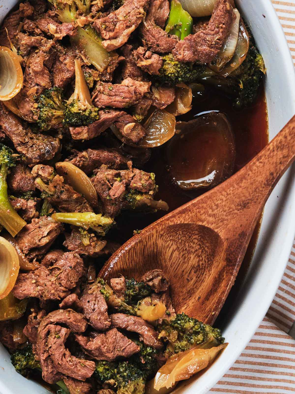A close-up image of a keto beef and broccoli stir-fry in a white dish with a wooden spoon.