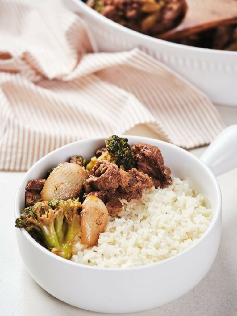A bowl of rice topped with keto beef and broccoli, with a cloth and another dish blurred in the background.