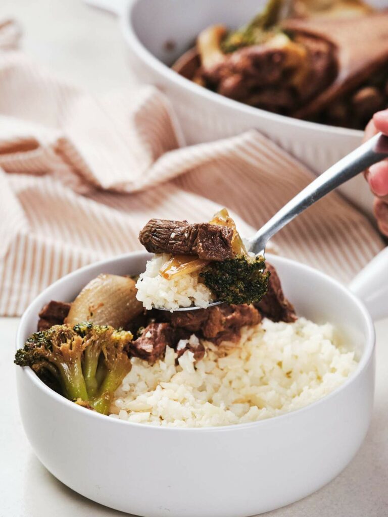 A bowl of keto beef and broccoli, with a fork lifting some food, set on a light-colored tablecloth.