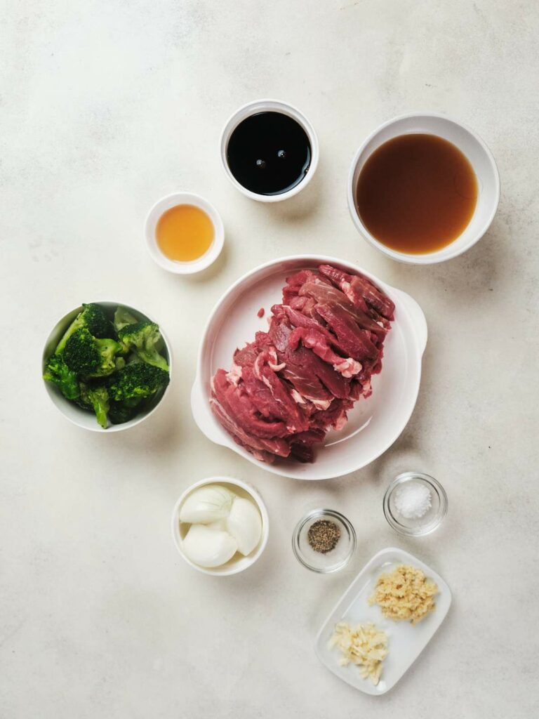 Ingredients for a keto beef and broccoli recipe neatly arranged on a table, including sliced raw beef, broccoli, garlic, onions, and various seasonings.